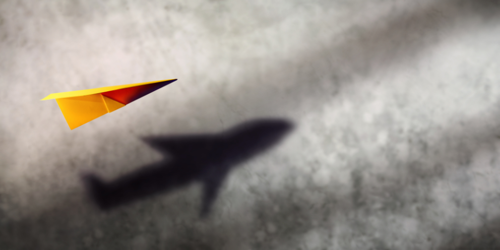 Yellow paper plane with a shadow of a bigger more detailed actual plane against grey background