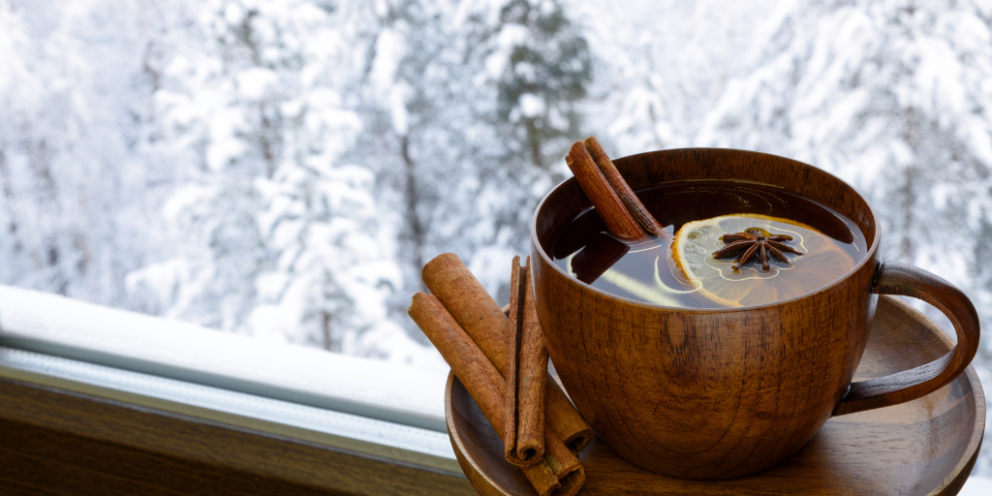 Brown mug filled with tea with sticks of cinnamon next to it on a windowsill looking over a snowed over landscape