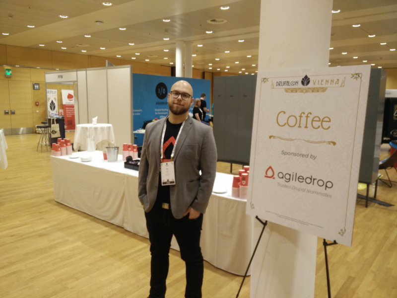 Agiledrop's then commercial director Iztok posing in front of coffee sponsors sign at DrupalCon Vienna 2017
