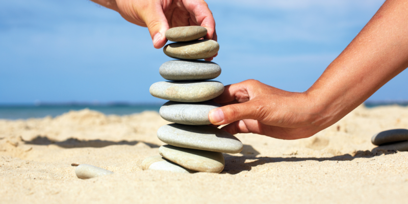 Two suntanned hands attempting to balance a pillar of rocks on a beach