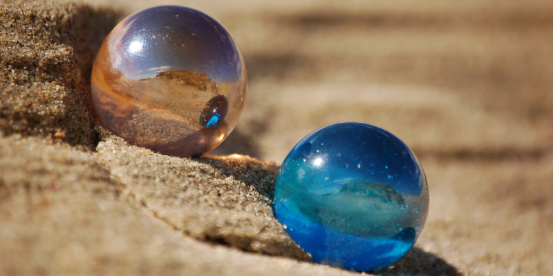 A orange and a blue marble sitting side by side on a rocky/sandy setting